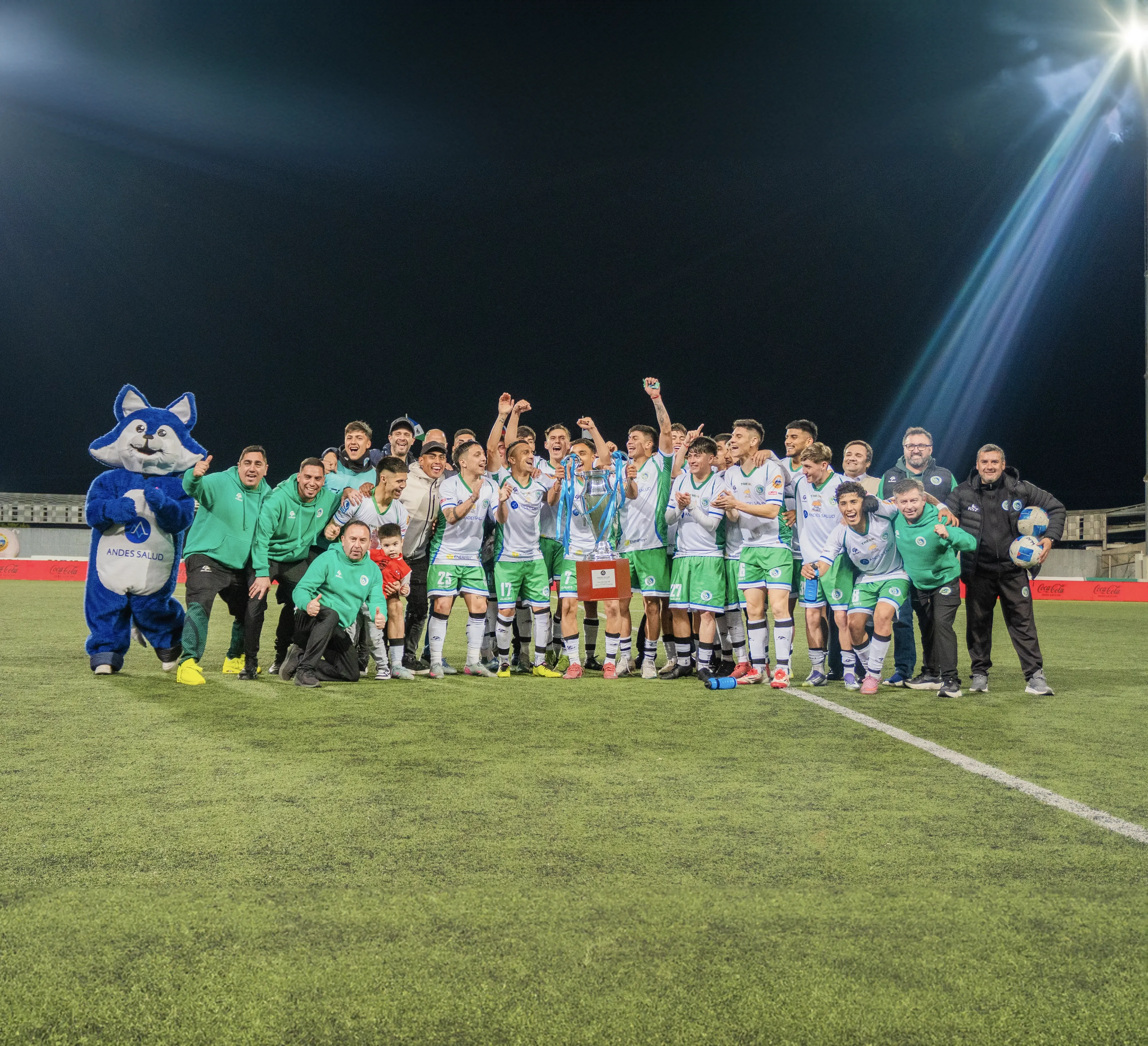 Equipo deportivo celebrando con un trofeo junto a la mascota de Andes Salud en una cancha iluminada.